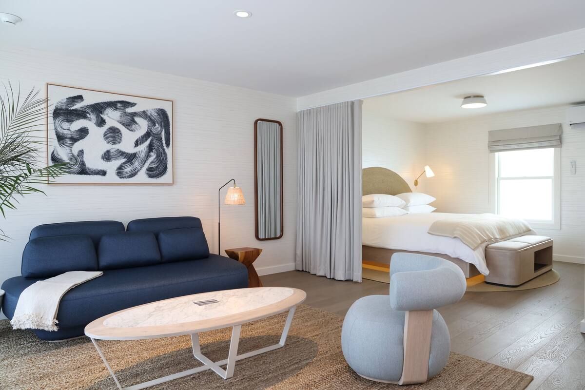 A modern hotel room with a blue sofa, a round coffee table, a large bed with a headboard, and a large mirror.