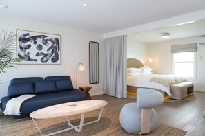 A modern hotel room with a blue sofa, a round coffee table, a large bed with a headboard, and a large mirror. - The Shoals Suites and Slips (Southold)