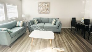 Una sala de estar con un sofá gris, una mesa de centro blanca y una mesa de comedor con sillas. Hay un cuadro en la pared y una lámpara sobre una mesita auxiliar.