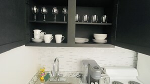 Una cocina con repisas empotradas que sostienen tazas blancas, platos y copas de vino. Hay una cafetera, un tostador y un lavamanos con grifo.