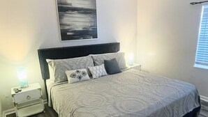 Un dormitorio con una cama grande, una mesita de noche, una lámpara de pared y un ventanal con persianas.