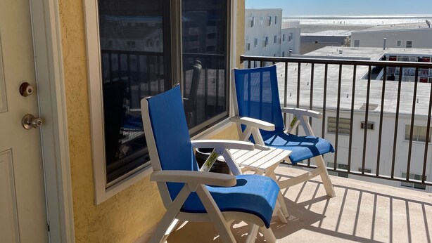 A balcony with two blue lounge chairs, a white door, and a large window offering a view of the beach and buildings.