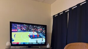 A living room with a flat-screen TV displaying a sports game, a wicker chair, a fireplace, and a dark curtain.