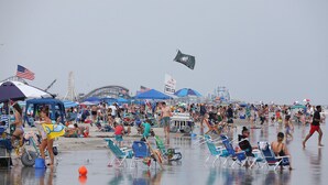 A crowded beach with numerous people, beach chairs, umbrellas, and a Ferris wheel in the background.