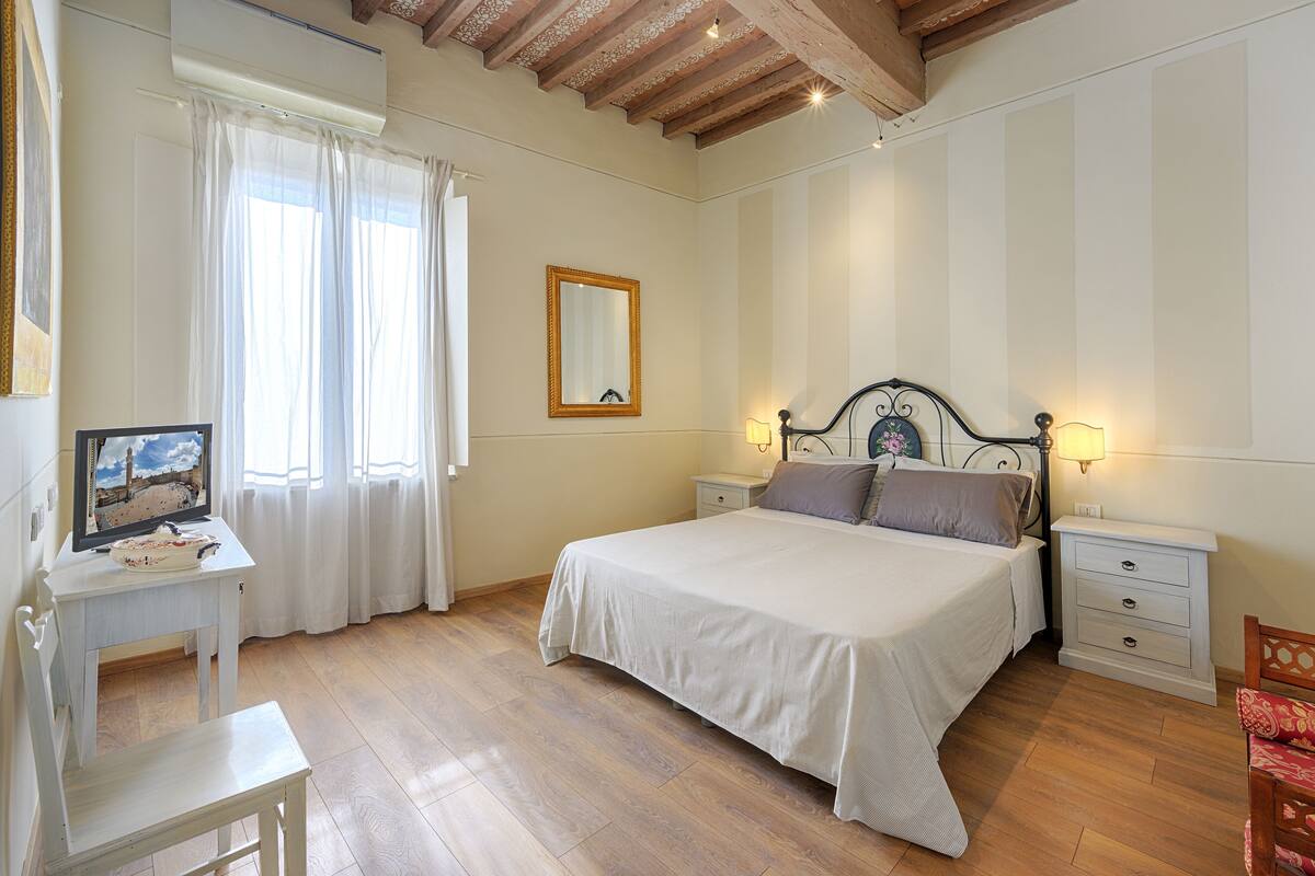 Une chambre à coucher comprenant un lit, des tables de chevet, une chaise, un bureau avec un ordinateur et une fenêtre avec des rideaux.