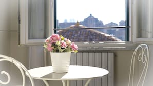 Una mesa blanca con un jarrón blanco que contiene flores rosas, ubicada frente a una ventana con vistas a un edificio y un tejado.