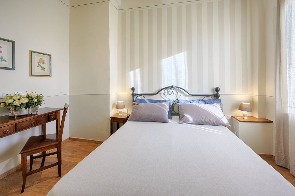 Une chambre à coucher bien rangée, avec un grand lit, deux tables de chevet avec des lampes, un bureau en bois avec une chaise et des tableaux encadrés accrochés au mur.