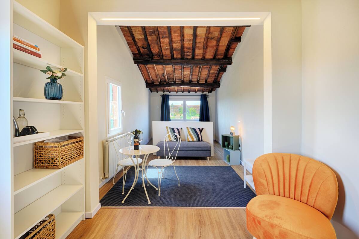 Une pièce chaleureuse avec un plafond en bois, un canapé agrémenté de coussins colorés, une petite table avec des chaises et une bibliothèque contenant divers objets.