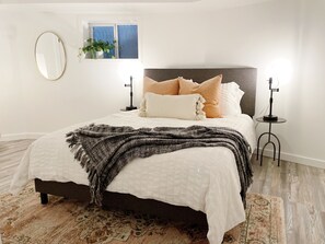 A neatly made bed with a white duvet, a dark headboard, and multiple pillows. There is a round mirror on the wall, a small potted plant, and a lamp on a side table.
