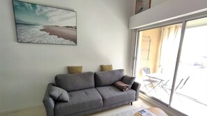 Un salon moderne avec un canapé gris, des coussins à motifs, une grande fenêtre et une petite table en bois sur laquelle repose un magazine.