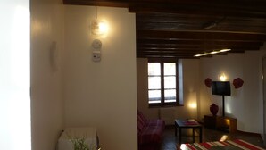 Uma sala de estar acolhedora, com um sofá, uma mesa pequena e uma televisão. Há uma janela com vista para o exterior e o teto possui vigas de madeira.