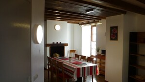 Uma sala de jantar com mesa posta para quatro pessoas, cadeiras de madeira, lareira e janela com iluminação natural.