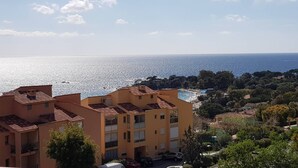 A coastal residential area with terracotta-roofed buildings, a clear view of the sea, and lush greenery.