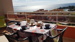 A balcony table set with a tablecloth, plates, bowls, and a pitcher, overlooking a coastal view with buildings and trees.