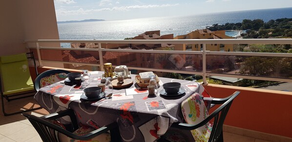 A balcony table set with a tablecloth, plates, bowls, and a pitcher, overlooking a coastal view with buildings and trees.