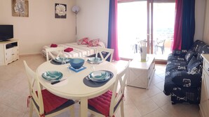 A well-lit room with a dining table set for two, a sofa, a TV, and a balcony with a view.