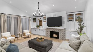 A modern living room with a grey sofa, a black ottoman, and a TV mounted above a fireplace. There are white cabinets with decorative baskets and a large window with sheer curtains. - Your Private Resort in SoCal Awaits (Long Beach)