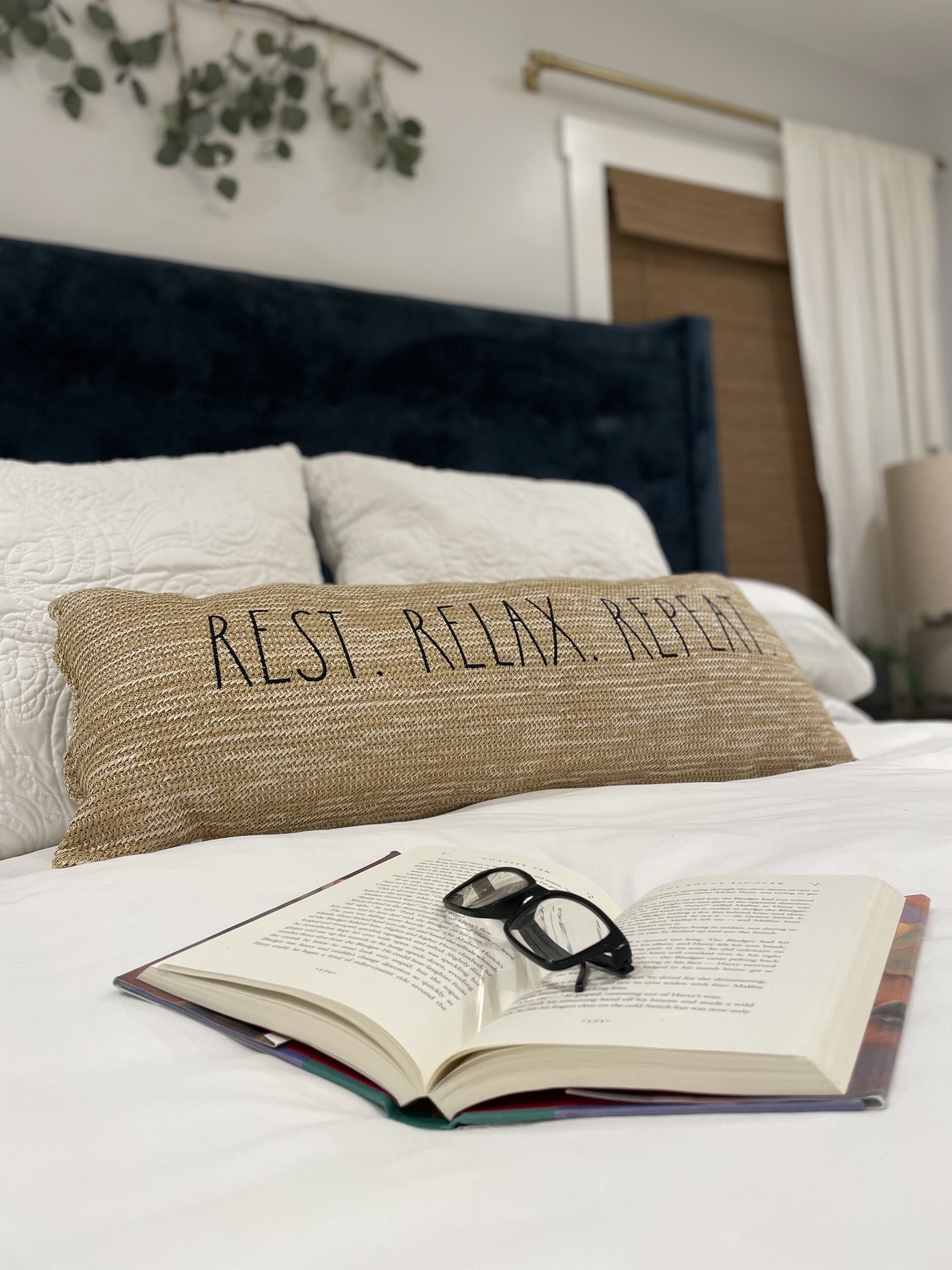 Een bed met een boek en een bril erop, een kussen met de tekst “RELAX RELAX RELAX”, en een plant die aan de muur hangt.