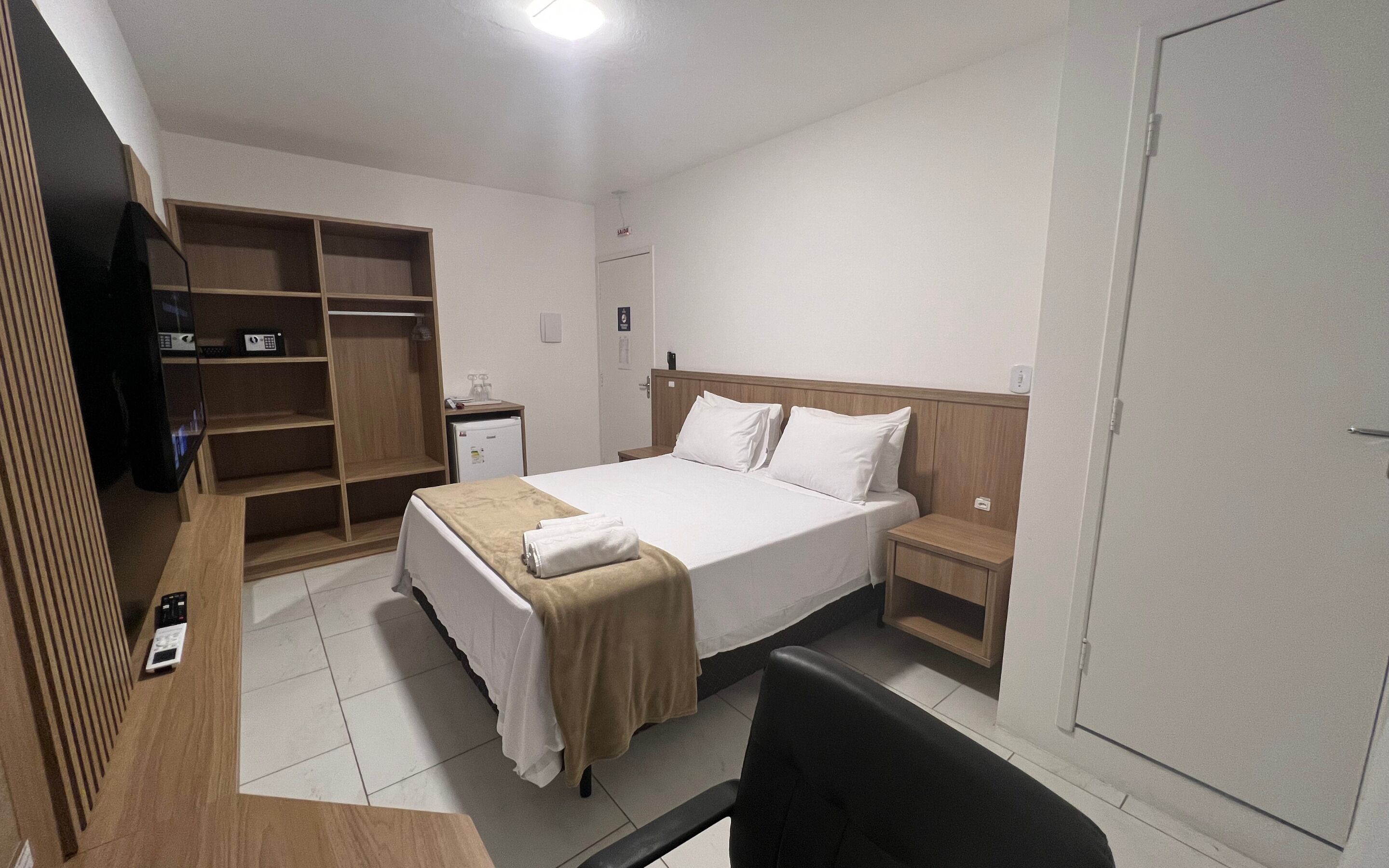 A hotel room with a bed, bedside tables, a desk with a chair, a wardrobe, and a television.