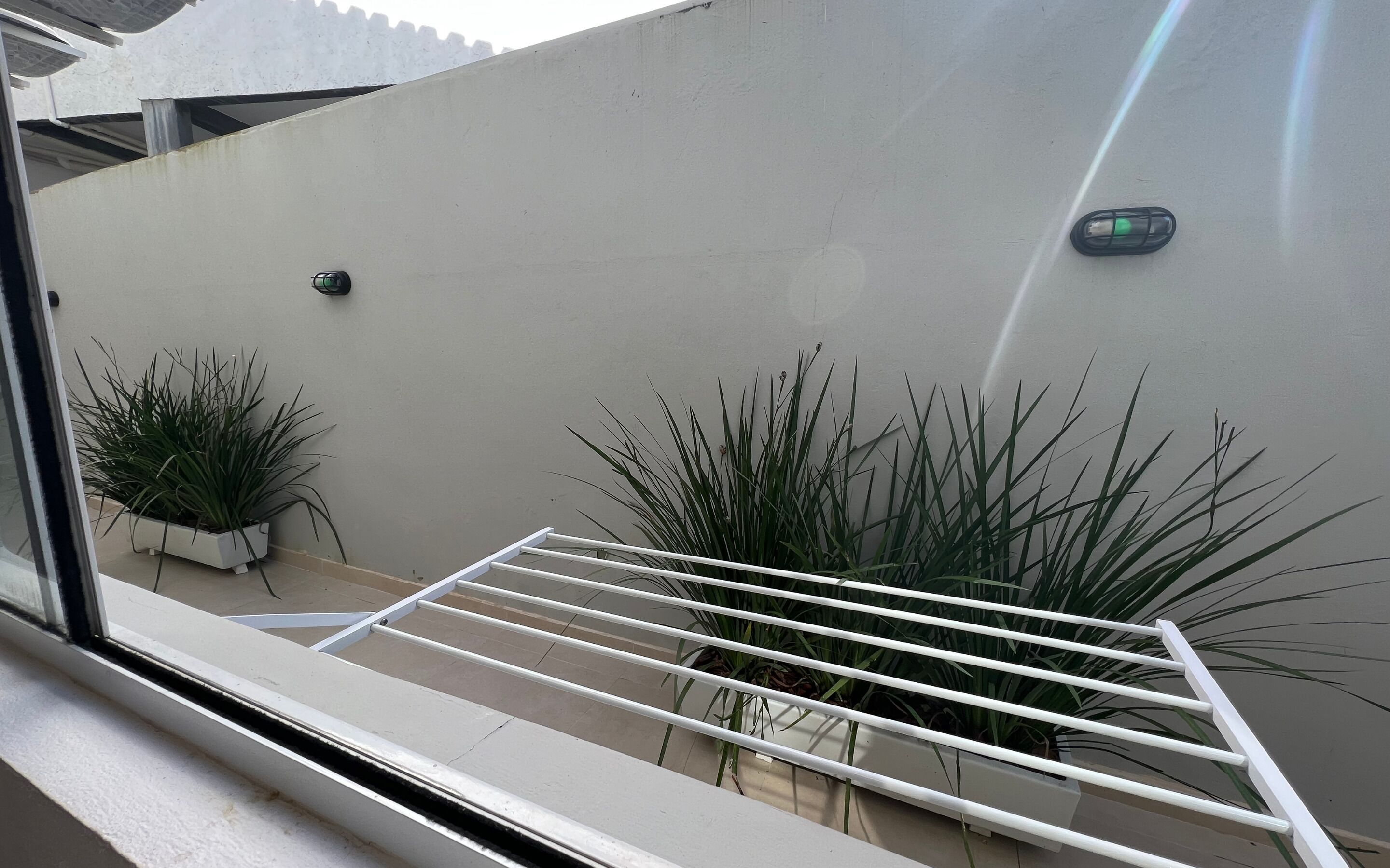 Una ventana con un estante metálico blanco, dos plantas en macetas y un aplique de pared.