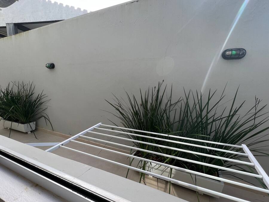Una ventana con un estante metálico blanco, dos plantas en macetas y un aplique de pared.