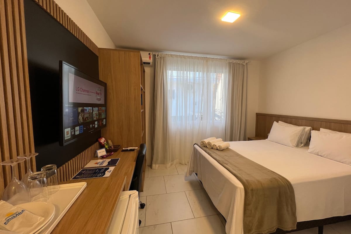 A hotel room with a bed, a desk with a TV displaying channels, a window with sheer curtains, and a wooden headboard.