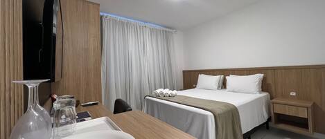 A hotel room with a bed, a desk, a TV, a nightstand, and a window with curtains.