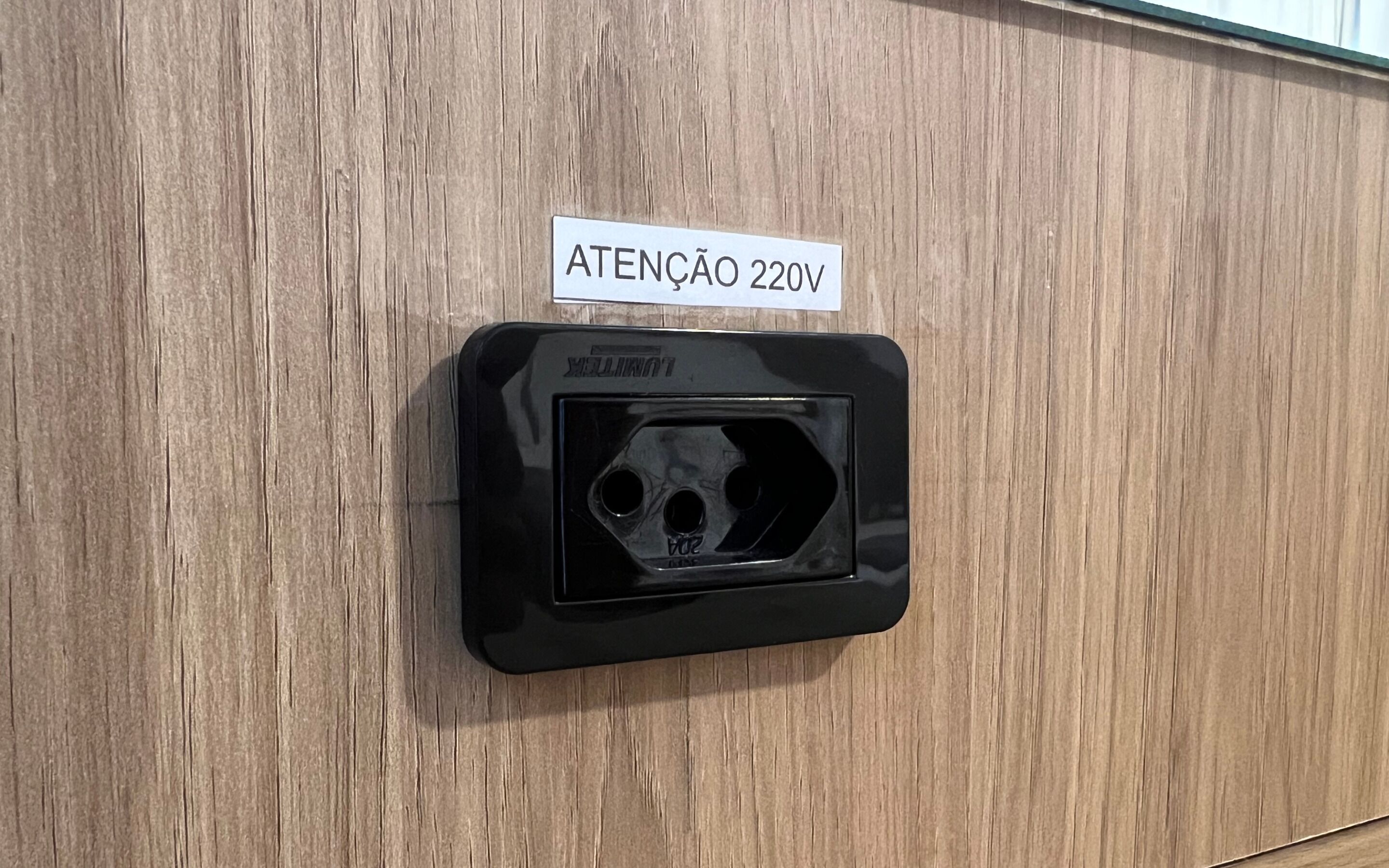 A black electrical outlet with a label reading "ATENÇÃO 220V" on a wooden wall.