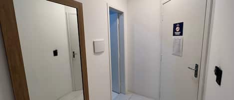 Un pasillo con paredes blancas, una puerta con marco de madera y una puerta con marco azul. Encima de la puerta azul hay un cartel que dice "SAIDA" y otro cartel que prohíbe fumar.