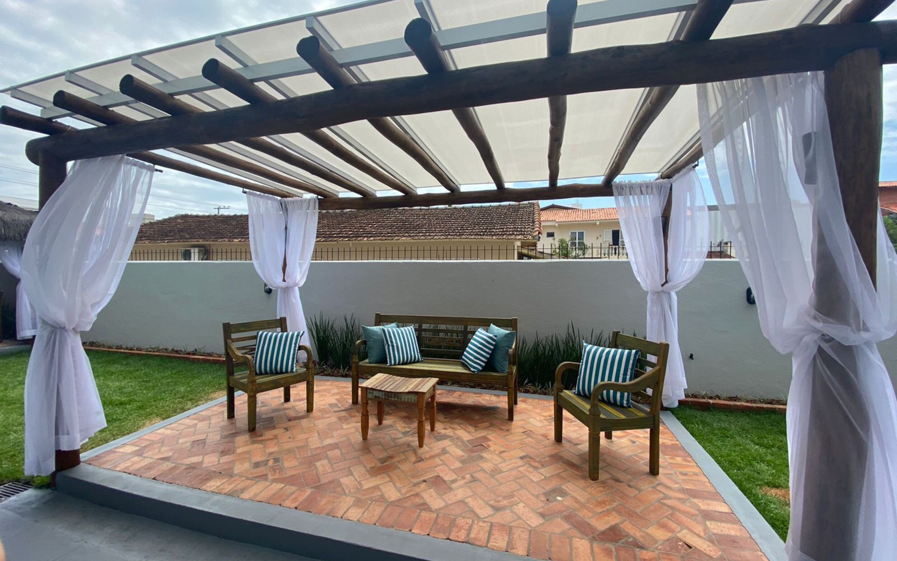 A covered patio area with a wooden bench and chairs, white curtains, and a brick floor.