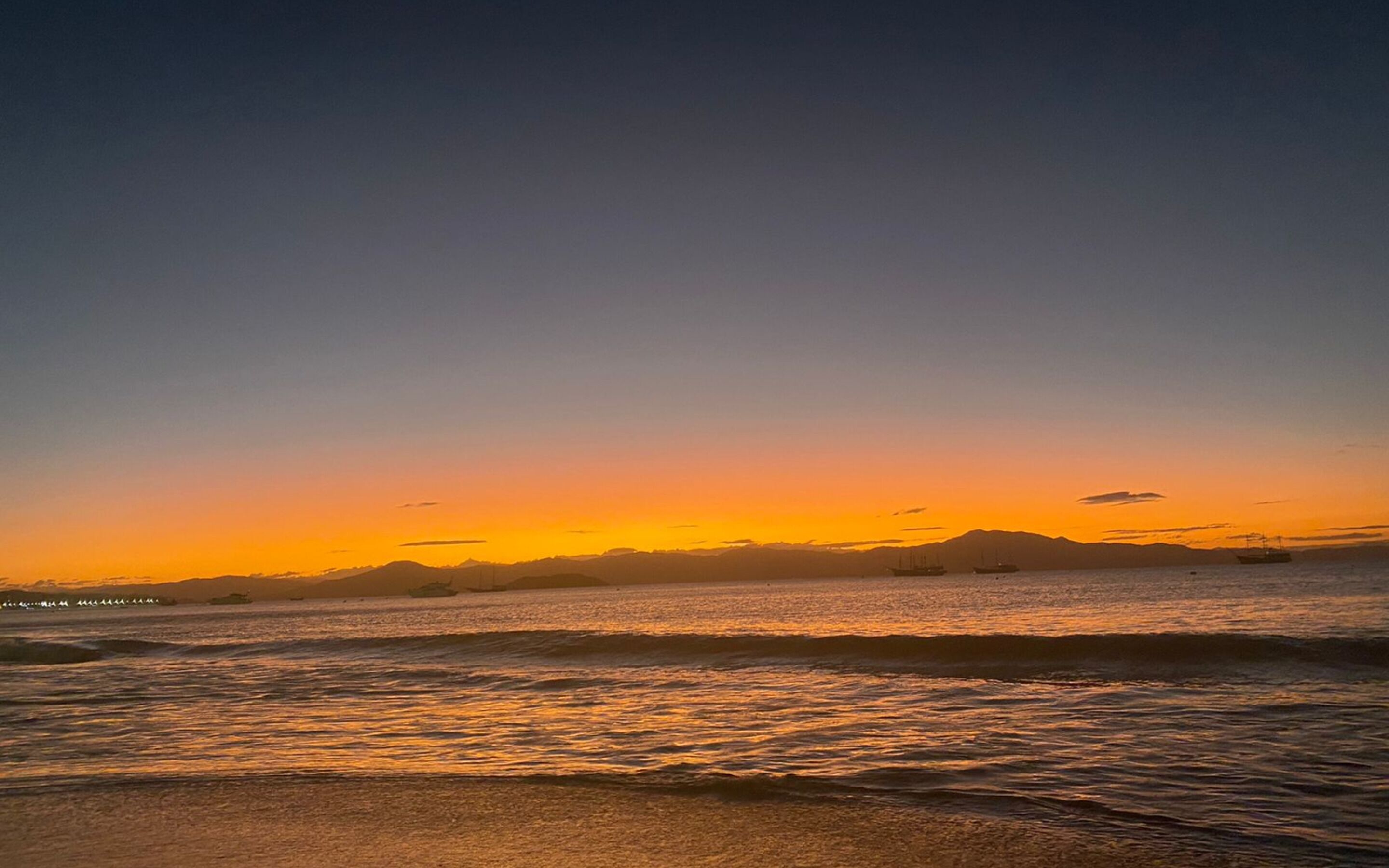 A beach at sunset with calm waves and distant mountains.