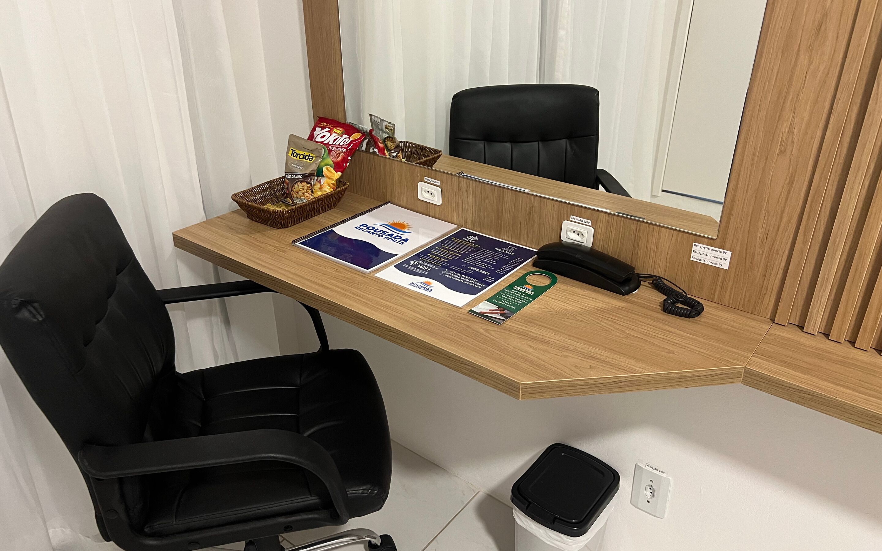 A hotel reception desk with a black office chair, a telephone, a basket of snacks, and informational brochures.