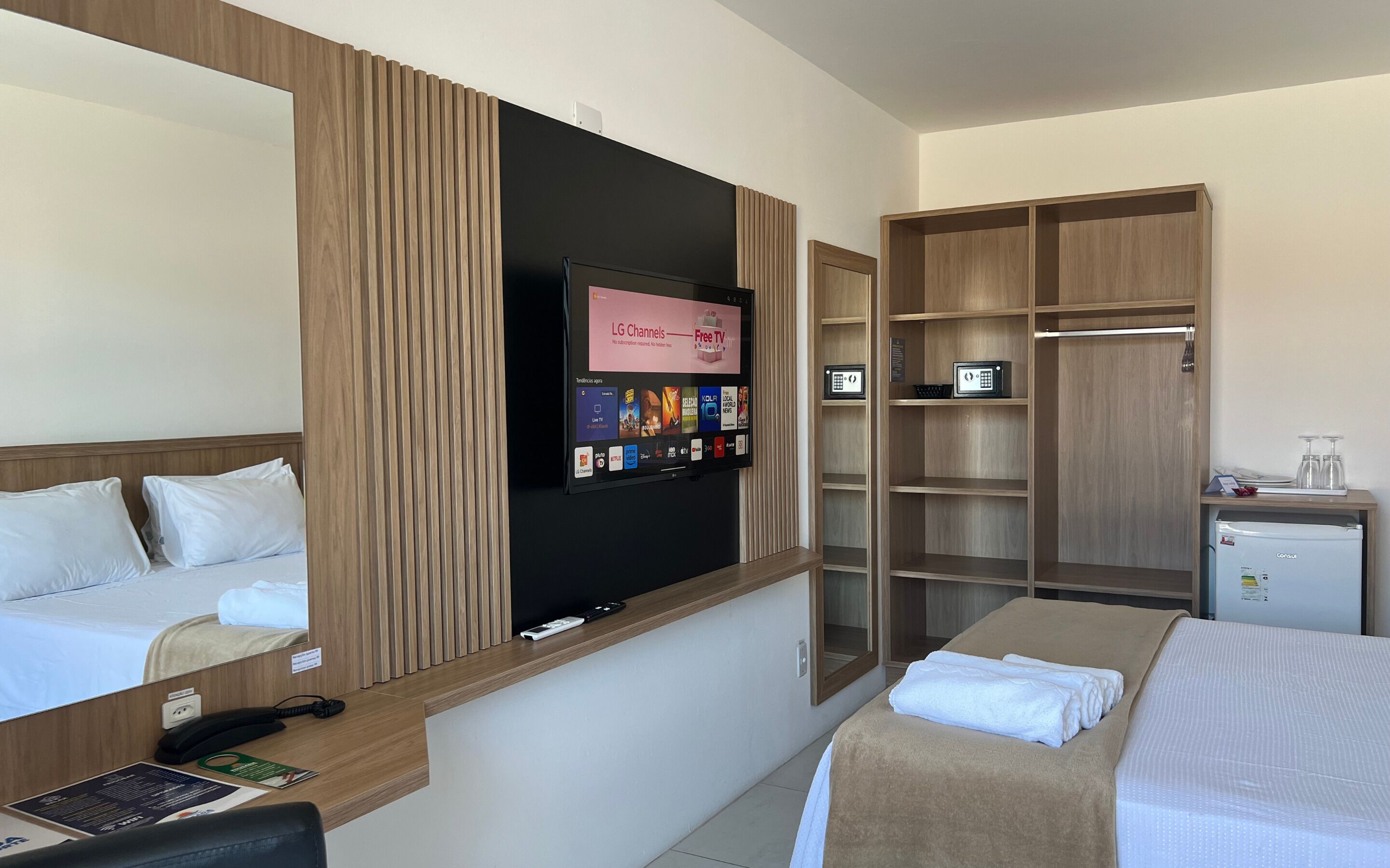 A hotel room with a bed, a desk with a phone, a TV mounted on the wall displaying channel options, a wardrobe, and a minibar.