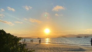 Escena de playa con el sol poniéndose sobre el océano, barcos en el agua y gente en la orilla.