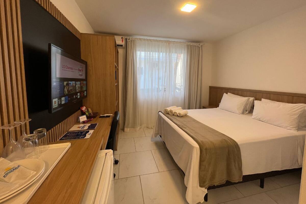 A hotel room with a bed, a desk with a flat-screen TV, a window with curtains, and a reception area with a counter and glasses.