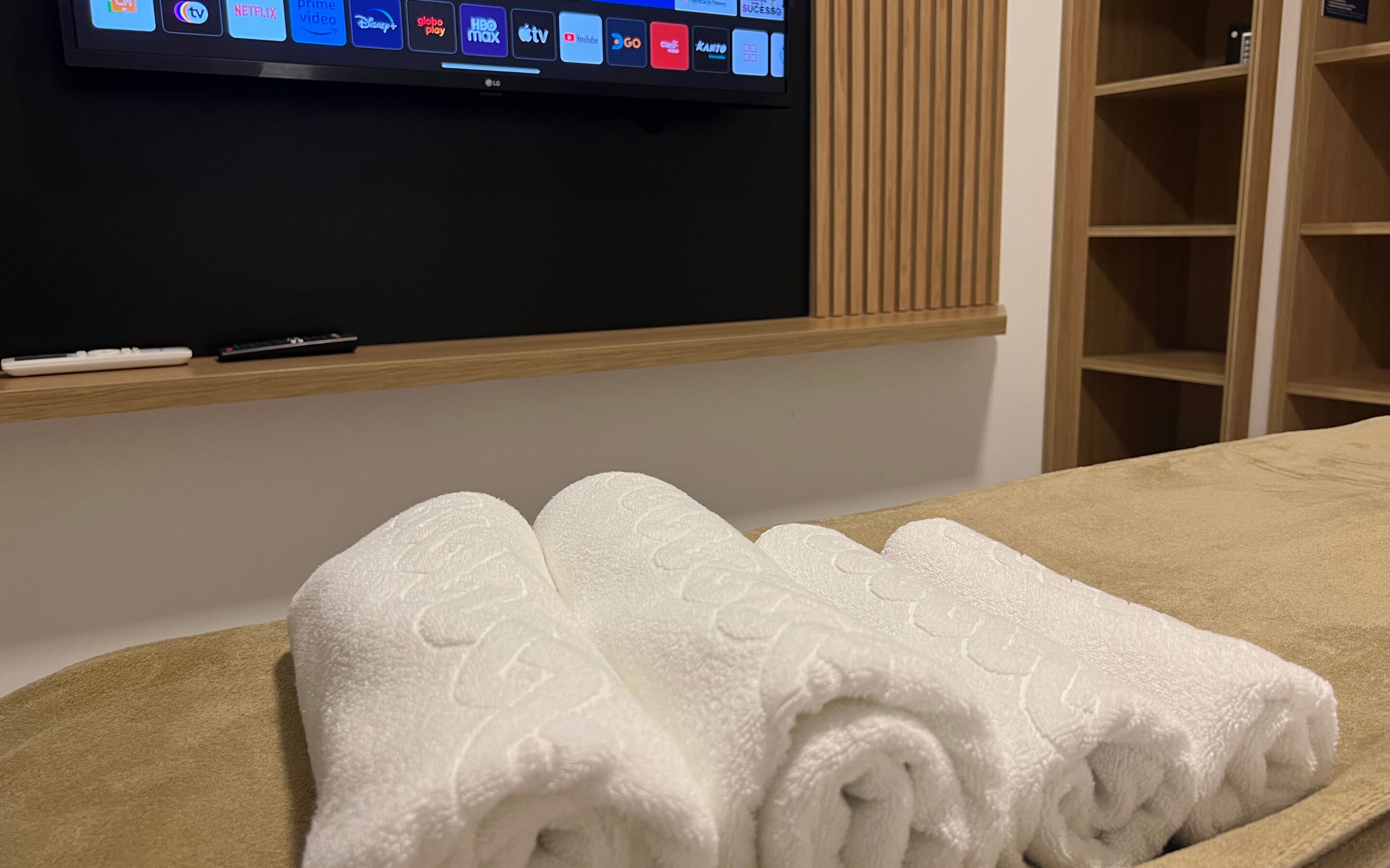 A hotel room with a flat-screen TV displaying various streaming services, a wooden shelf with a remote control, and a neatly folded white towel on the bed.