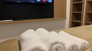 A hotel room with a flat-screen TV displaying various streaming services, a wooden shelf with a remote control, and a neatly folded white towel on the bed.