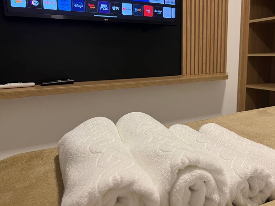 A hotel room with a flat-screen TV displaying various streaming services, a wooden shelf with a remote control, and a neatly folded white towel on the bed.