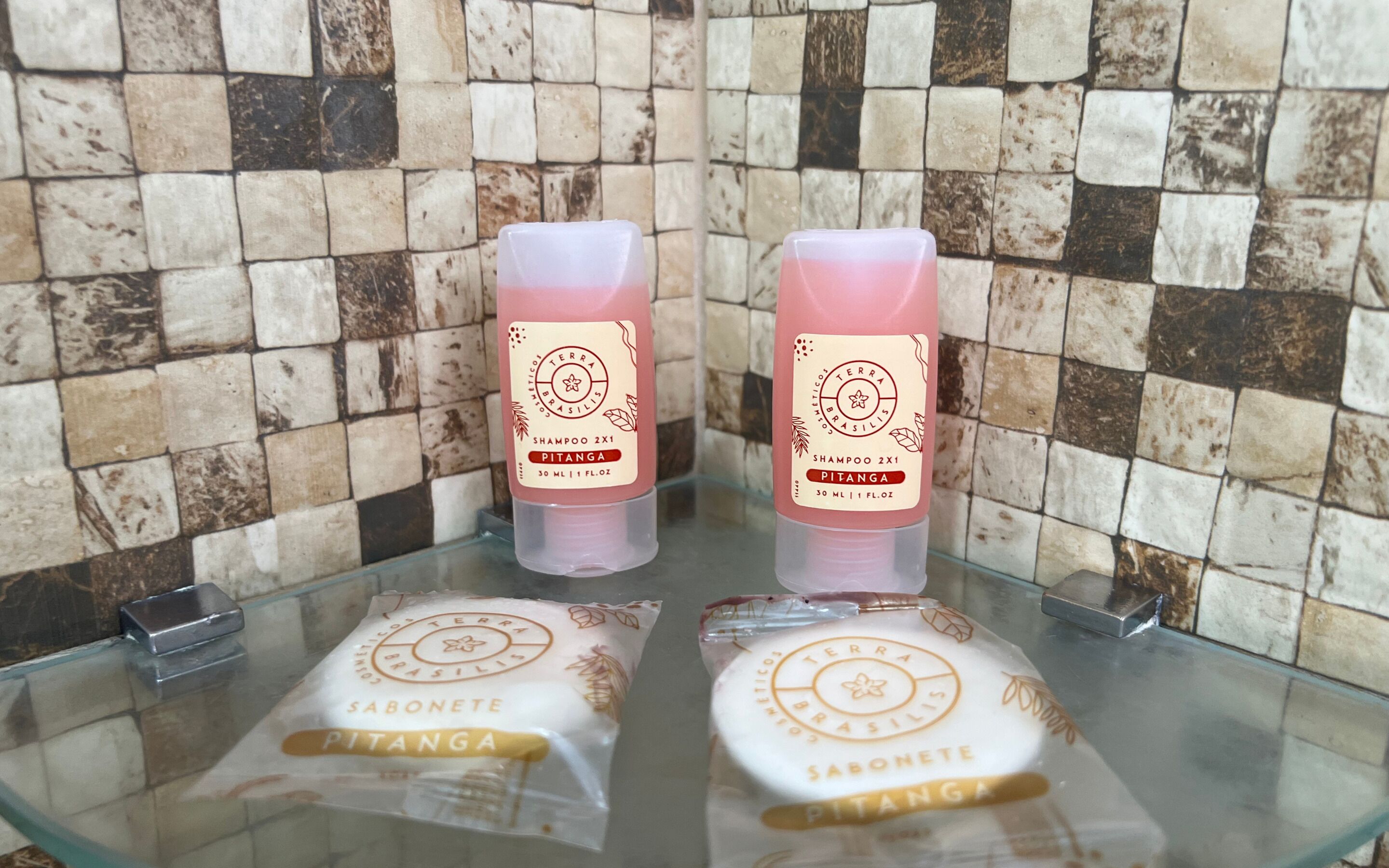 Two bottles of shampoo and two sachets of soap on a glass shelf against a tiled wall.