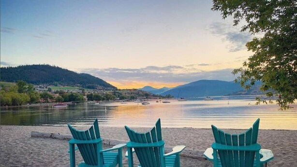 Een strand met drie turquoise Adirondack stoelen, gericht op een rustig meer met boten en een zonsondergang.
