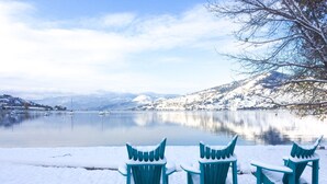 Drie met sneeuw bedekte Adirondack-stoelen met uitzicht op een rustig meer en een bergachtige achtergrond.