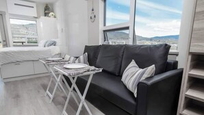 Een moderne woonkamer met een zwarte bank, een klein wit tafeltje met een bord, en een stadszicht door grote ramen.