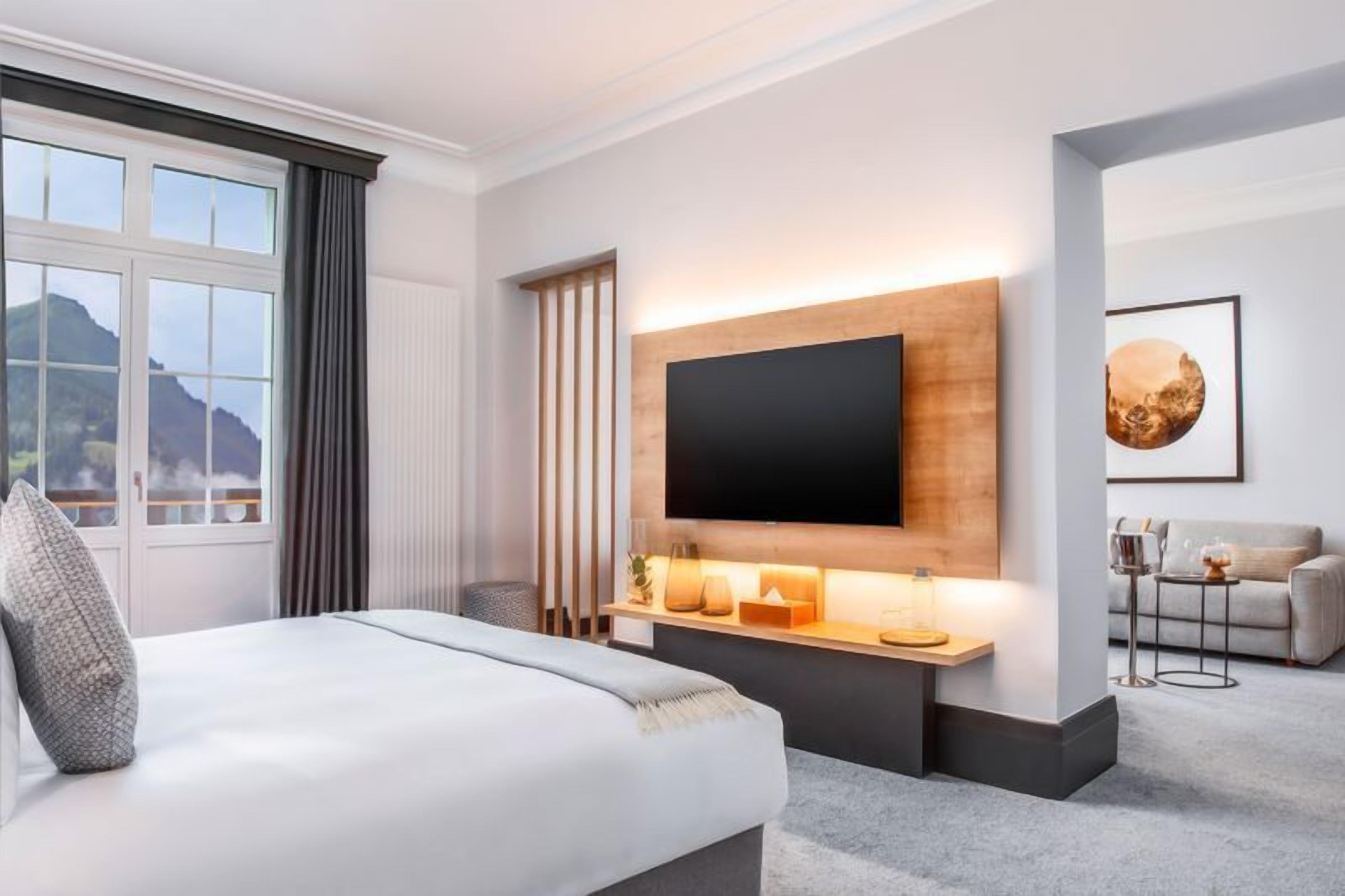 A hotel room with a large bed, a mounted TV, a wooden shelf with decorative items, a sofa, and a view of mountains through the window.