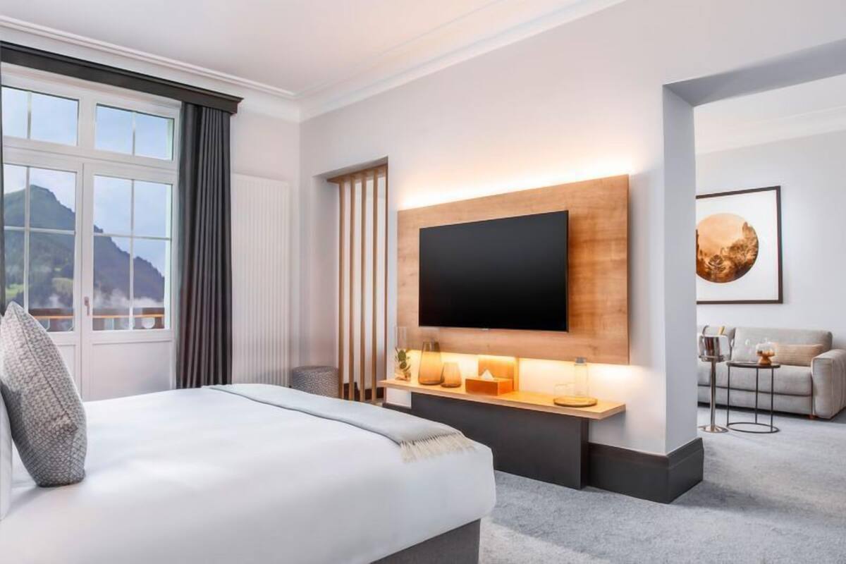 A hotel room with a large bed, a mounted TV, a wooden shelf with decorative items, a sofa, and a view of mountains through the window.
