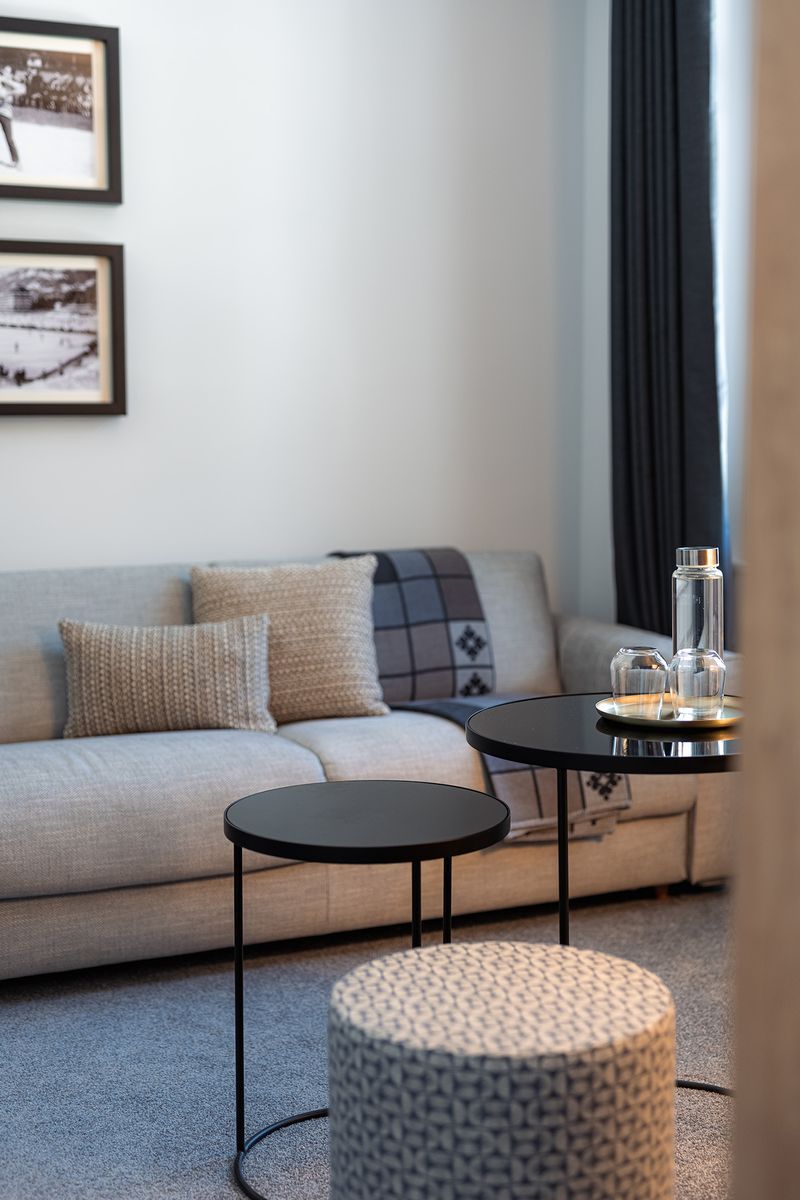 A modern living room with a sofa, a round coffee table, and a small ottoman. There are framed pictures on the wall and a window with curtains.