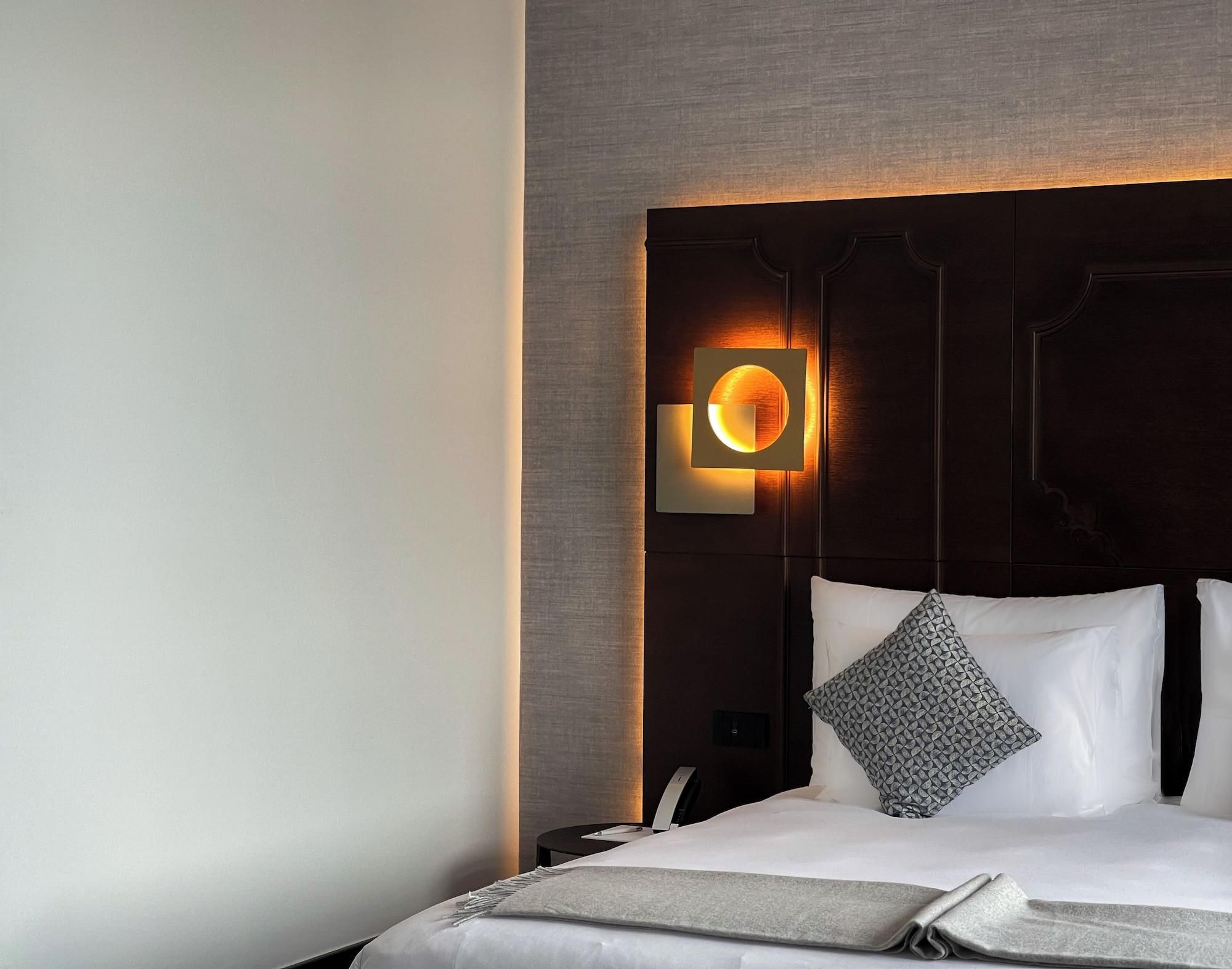 Une chambre d’hôtel au design moderne, dotée d’un grand lit avec du linge de lit blanc, d’une tête de lit sombre et d’un luminaire mural original.