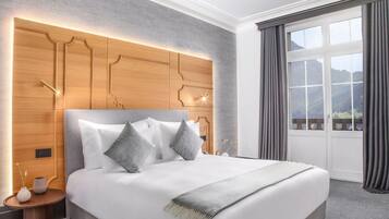 Ein modernes Hotelzimmer mit einem großen Bett, einem hölzernen Kopfteil mit integrierter Beleuchtung, einem Nachttisch und Blick auf Berge durch ein großes Fenster mit Vorhängen.