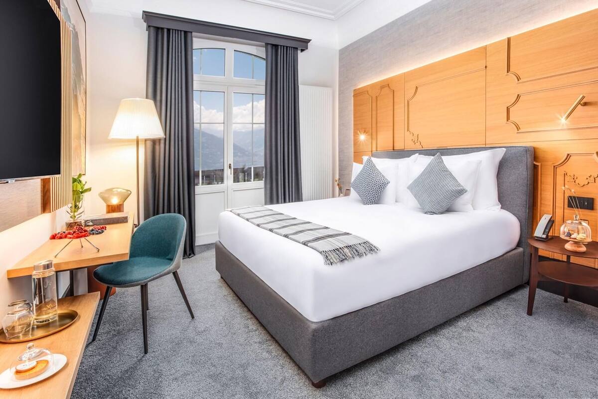 A hotel room with a bed, a desk, a chair, a TV, a window with curtains, and a wooden headboard.