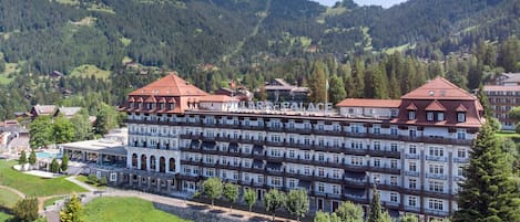 A large hotel with multiple floors and a red roof, surrounded by lush greenery and mountains.