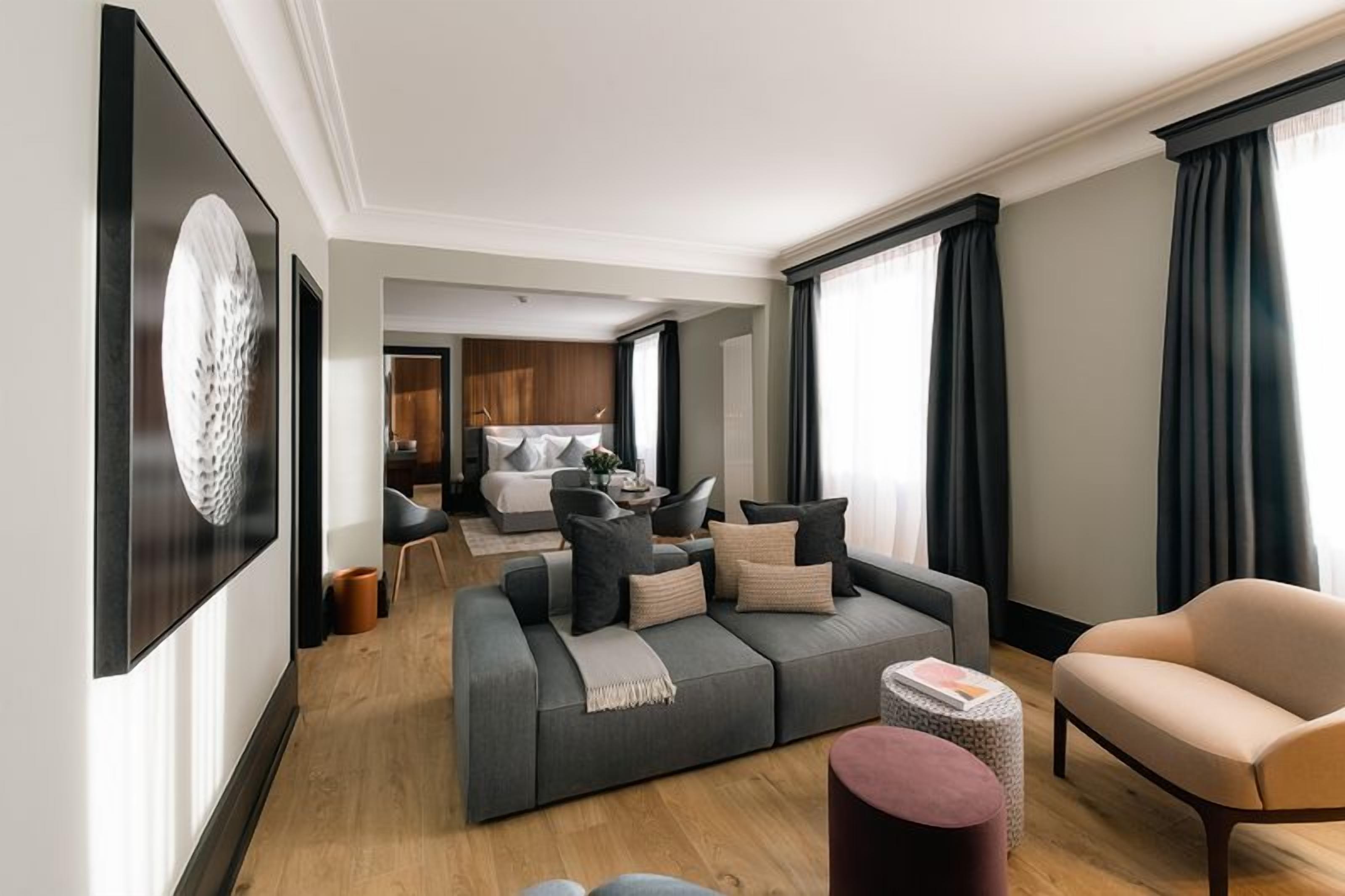 A modern hotel room with a grey sofa, a beige armchair, and a small round table. There is a large window with dark curtains, a framed picture on the wall, and a wooden floor.